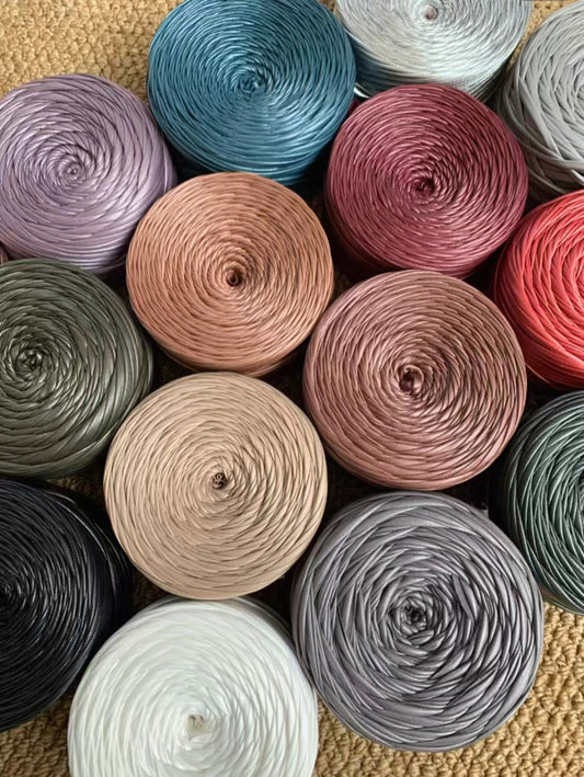 A vibrant flat lay arrangement of multiple cakes of premium matte satin faux leather ribbon yarn. The various cakes display a rich and harmonious color palette, including muted metallics, jewel tones, and neutrals such as a dusty rose, sage green, deep teal, copper bronze, slate gray, olive, matte black, pure white, and soft silver gray, showcasing the smooth, durable texture and uniform color.