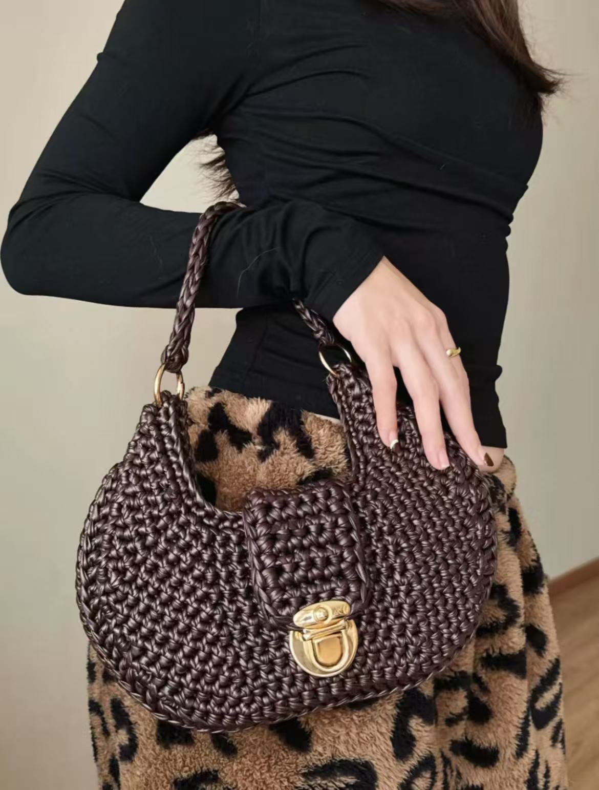 A person wearing a black long-sleeved top and a brown leopard print skirt is shown holding a luxurious, hand-crocheted shoulder bag made from deep chocolate brown matte faux leather yarn. The bag is a structured hobo style, featuring a detailed, robust basketweave pattern, an ornate gold-tone twist lock closure on the central flap, and matching gold-tone hardware on the sturdy, braided leather-yarn handle, illustrating the refined texture and shape-holding capability of the finished product.