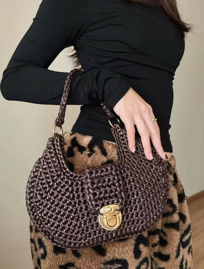 A person wearing a black long-sleeved top and a brown leopard print skirt is shown holding a luxurious, hand-crocheted shoulder bag made from deep chocolate brown matte faux leather yarn. The bag is a structured hobo style, featuring a detailed, robust basketweave pattern, an ornate gold-tone twist lock closure on the central flap, and matching gold-tone hardware on the sturdy, braided leather-yarn handle, illustrating the refined texture and shape-holding capability of the finished product.